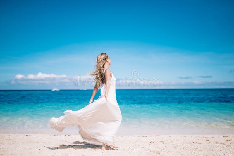 Beautiful blonde long hair bride in long white dress running on the white sand beach. Tropical turquois sea on the background.