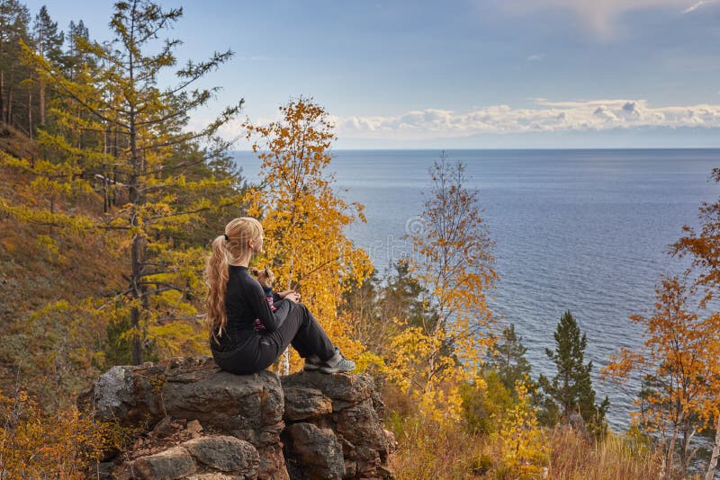 The beautiful blonde sits on the rock with a dog and admires a view of the autumn lake