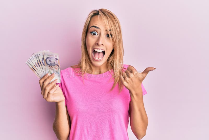 Beautiful blonde woman holding 2 singapore dollars banknotes pointing thumb up to the side smiling happy with open mouth