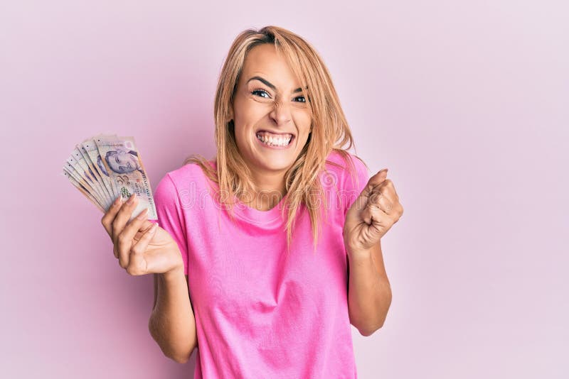 Beautiful blonde woman holding 2 singapore dollars banknotes screaming proud, celebrating victory and success very excited with