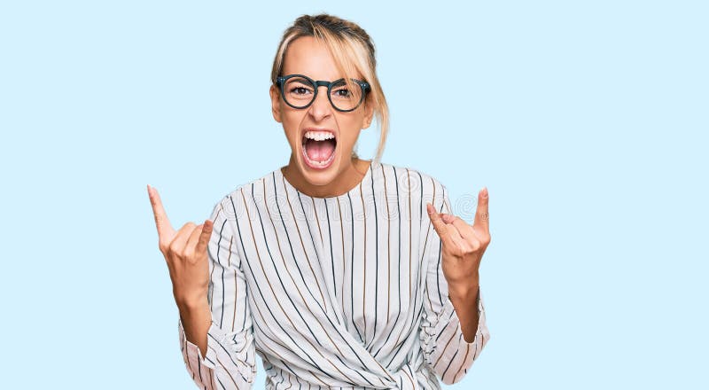 Beautiful blonde woman wearing business shirt and glasses shouting with crazy expression doing rock symbol with hands up