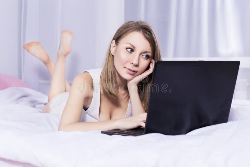 Beautiful blonde woman working on a laptop in her bed in pajamas, smiling