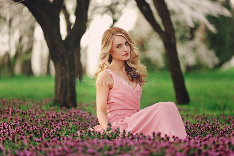 Beautiful blonde young woman in colorful flowers. girl with make-up and hairstyle in pink dress in blossoming spring park. woman`