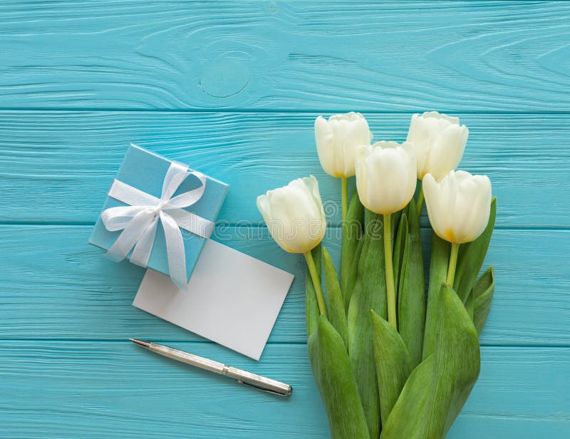 Beautiful bouquet of white tulips and blue gift box, empty card, pen on the blue wooden background