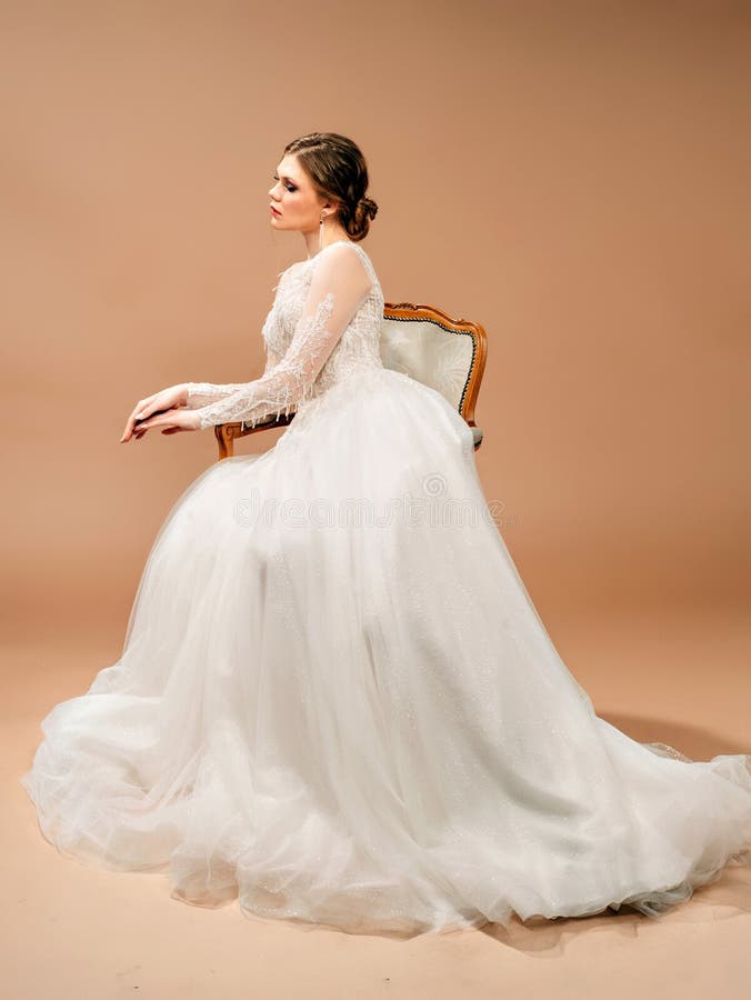 Beautiful bride in white wedding dress sitting in a vintage chair. Studio portrait with brown background.