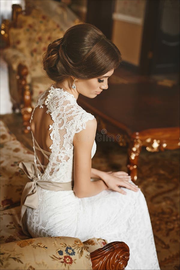 Beautiful bride, young model brunette woman, in stylish wedding dress with naked back sits on the vintage sofa and posing in luxur