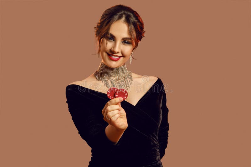 Brunette girl in black dress and shiny jewelry. She smiling, showing two red chips, posing on brown background. Poker