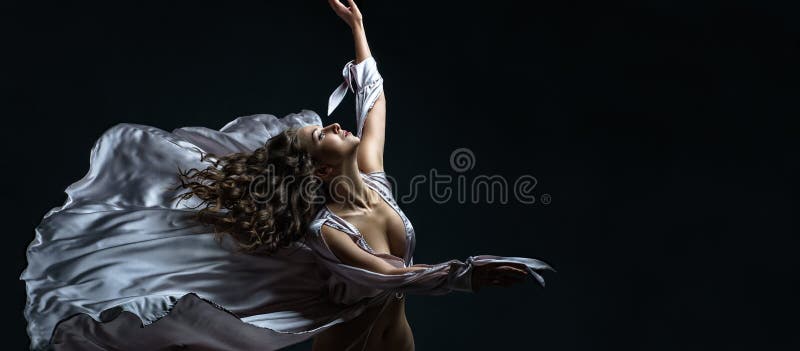 Beautiful brunette girl with curly hair in the darkness and light in silver satin flying dress awesome poses in dance