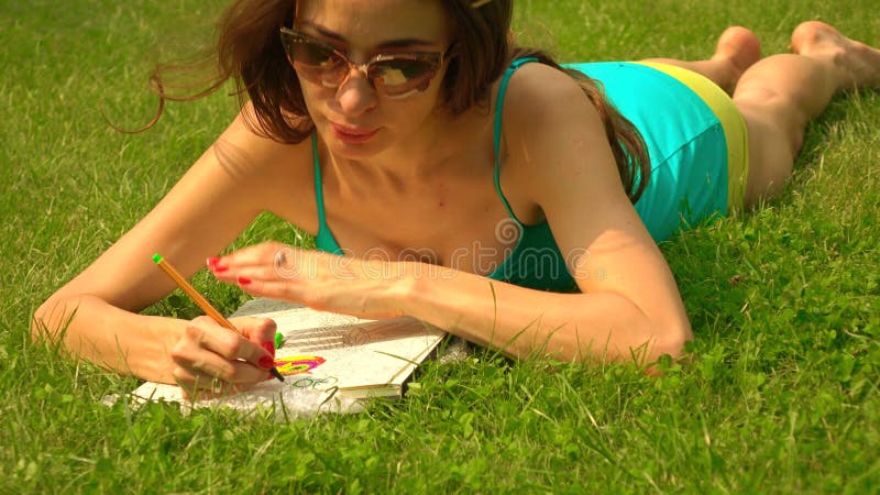 Beautiful brunette girl in sunglasses laying on the grass and colorizing. 4K steadicam shot, vivid colors