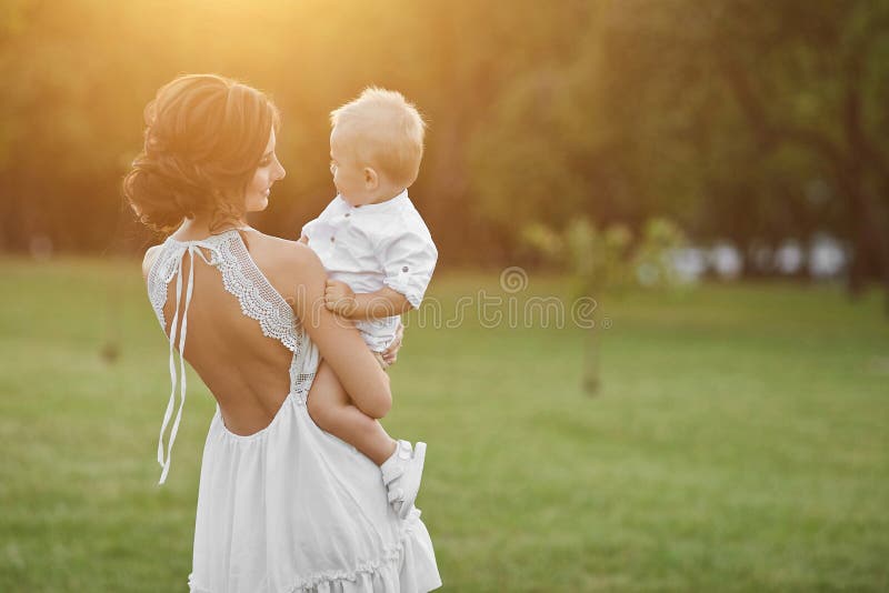 Beautiful brunette model girl, young mom in short white stylish dress holding on her hands her cute happy baby boy and stock photography Beautiful brunette model girl, young mom in short white stylish dress holding on her hands her cute happy baby boy and stock photography