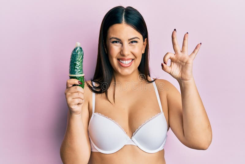 Beautiful Brunette Woman Holding Condom On Cucumber For Sex Education Doing Ok Sign With Fingers