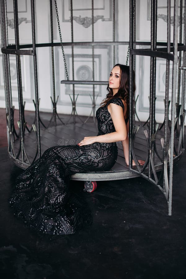Beautiful brunette woman wearing shiny black dress posing in cage like bird.