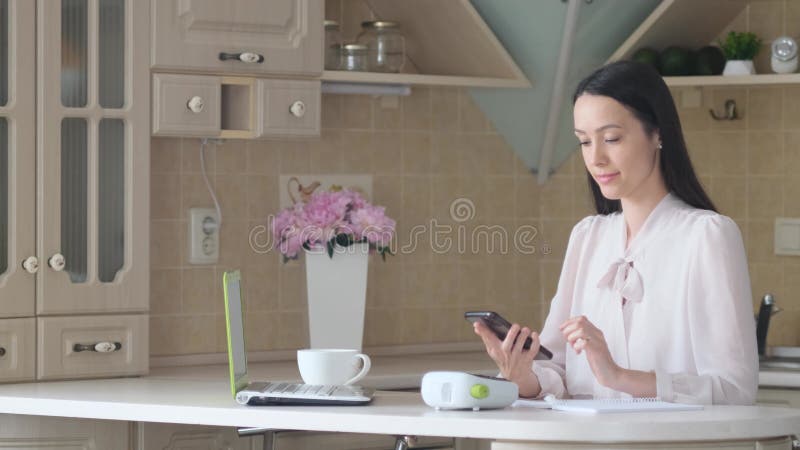 Beautiful brunette working on a laptop computer and using a mobile phone in the kitchen. stock video footage