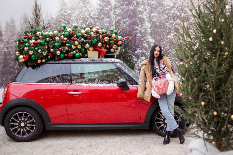 Beautiful young girl is holding gift box near red car with decorated xmas tree on the roof, holiday and new years