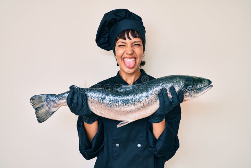 Beautiful brunettte woman professional chef holding fresh salmon fish sticking tongue out happy with funny expression stock image