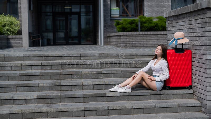 A beautiful caucasian woman wearing in a hat and shorts is sitting on the stairs with a large red suitcase. Smiling girl