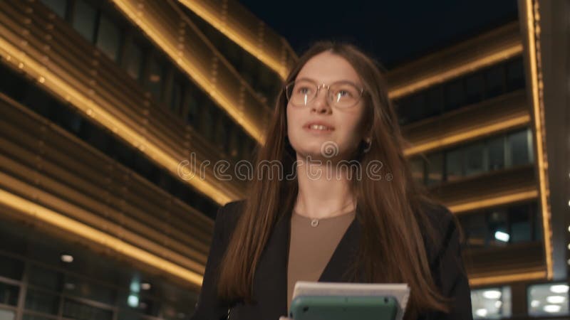 A happy businesswoman is walking in the evening after work, against the backdrop of an illuminated office building.