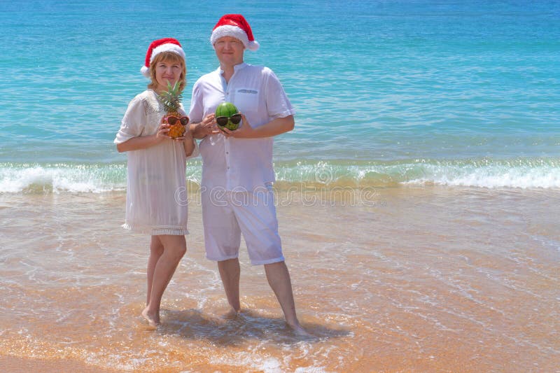 Beautiful couple in love having fun in the waves dressed in red Santa Claus hats with fruits in hand. Christmas on the tropical royalty free stock images Beautiful couple in love having fun in the waves dressed in red Santa Claus hats with fruits in hand. Christmas on the tropical royalty free stock images