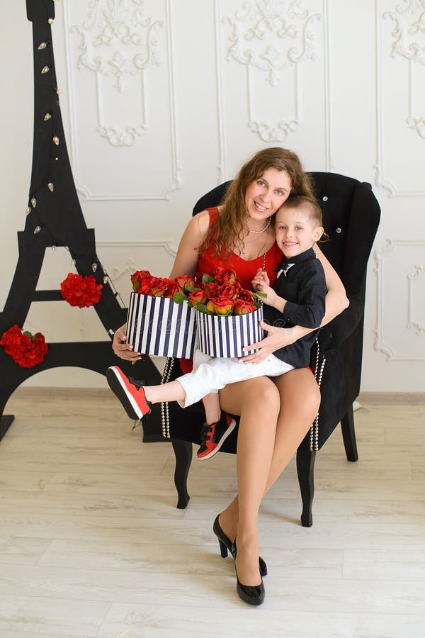 Curly-haired woman in red dress is sitting in black chair with small son in her arms. in her hands boxes with flowers