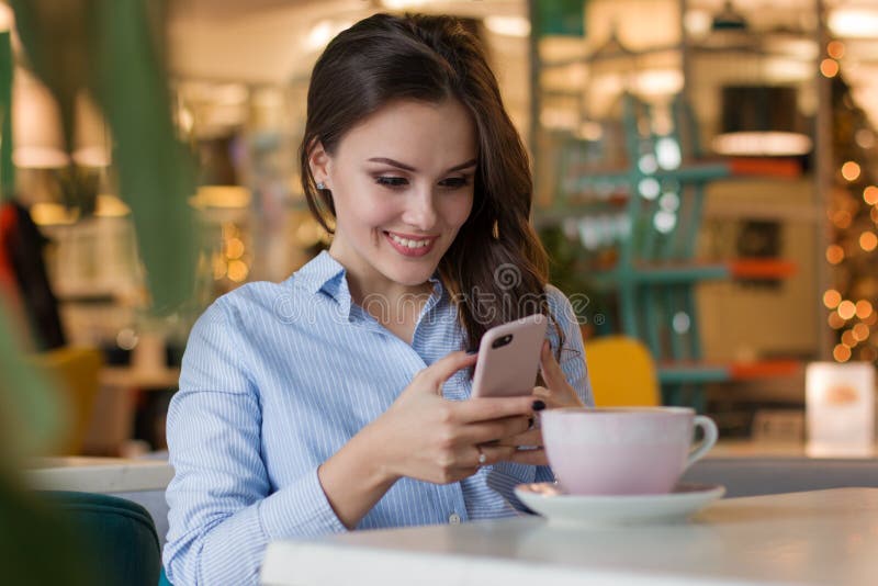 Beautiful cute caucasian young woman in the cafe, using mobile phone and drinking coffee smiling