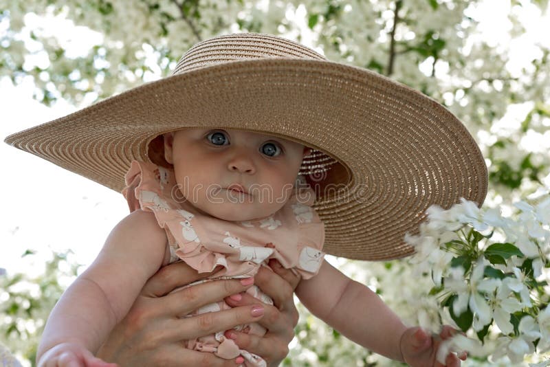 Beautiful cute infant in a big straw hat and with big blue eyes. Portrait of adorable baby on a spring blooming apple trees