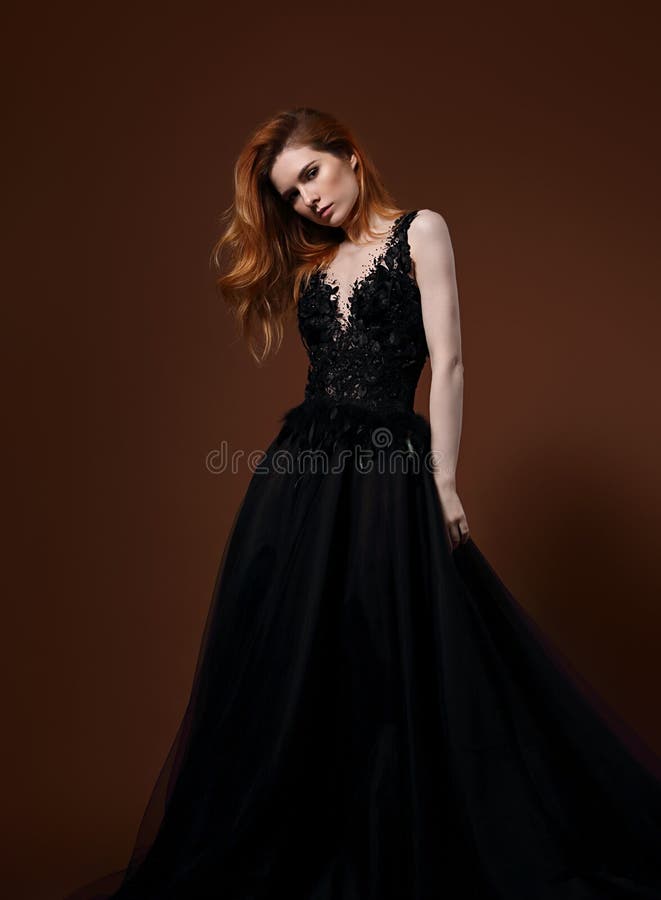 Beautiful elegant young model with bright redhead hairstyle posing in fashion chic black wedding dress with long skirt on studio