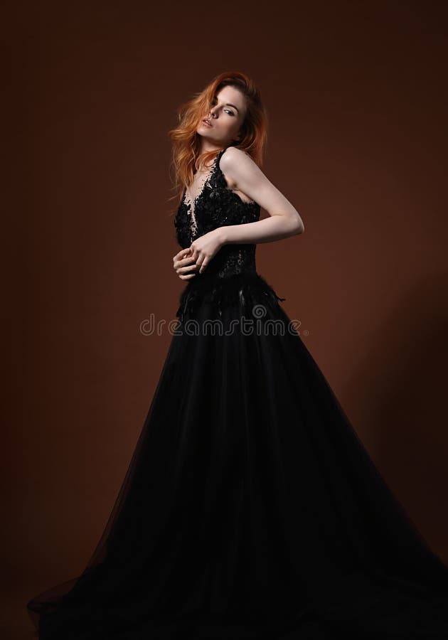 Beautiful elegant young model with bright redhead hairstyle posing in fashion chic black wedding dress with long skirt on studio