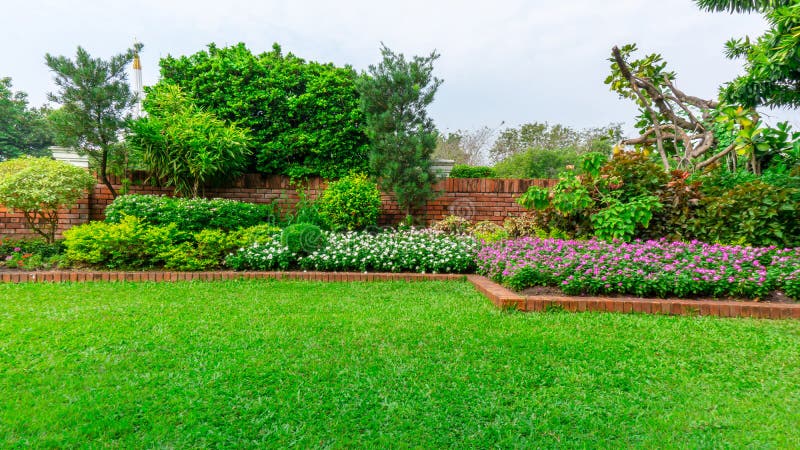 Beautiful English cottage garden, colorful flowering plant on smooth green grass lawn and group of evergreen trees in good care maintenance landscaping of a public park under white sky