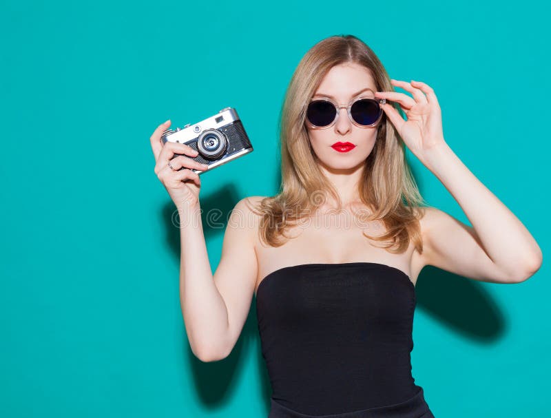 Beautiful fashionable girl posing and holding a vintage camera in black dress and sunglasses on the green background in the studio. Gorgeous Woman Portrait. Stylish Haircut and Makeup. Glamour Girl.