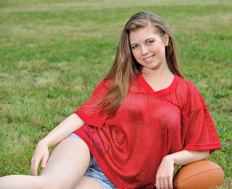 young Caucasian woman in red mesh football jersey and shorts - sitting in grass and on a football - smiling. young Caucasian woman in red mesh football jersey and shorts - sitting in grass and on a football - smiling