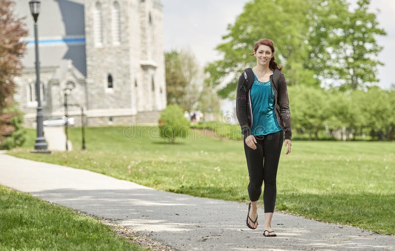 Beautiful female student, with red hair, wearing backpack on campus - walking down path. Beautiful female student, with red hair, wearing backpack on campus - walking down path