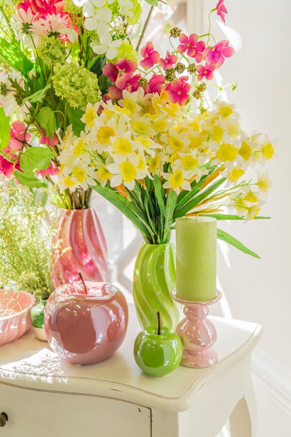 Beautiful flowers in green and pink vases, Candles and glass apples on white wooden  commode. Spring interior decoration.