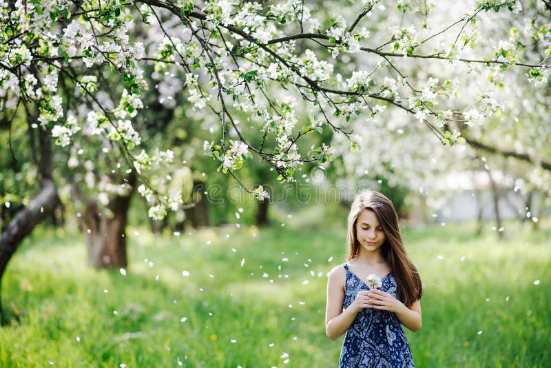 Beautiful girl in a blue dress in a blooming apple orchard. Allergy Season. Snow from flower petals