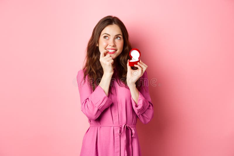 Beautiful girl dream of her wedding day, looking thoughtful up, showing engagement ring in red box and smiling, standing