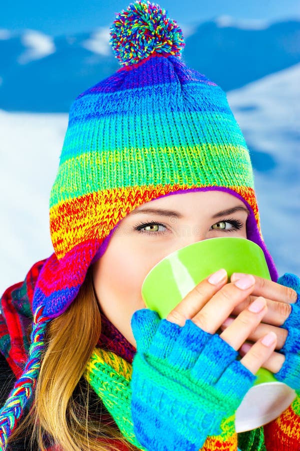 Beautiful girl drinking hot chocolate outdoor in winter, happy female teen wearing colorful hat, young pretty woman face, holding cup in hands