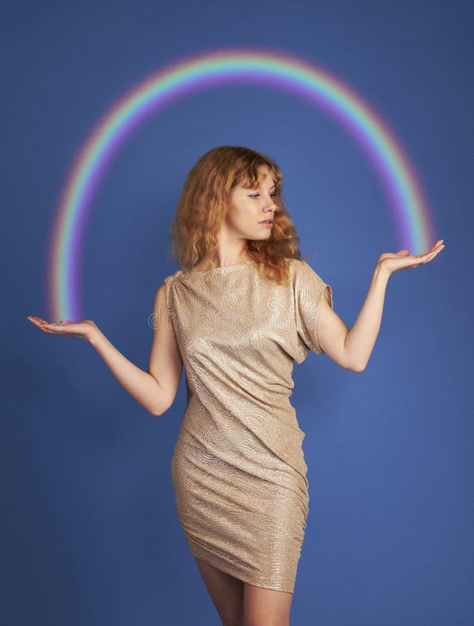 Girl with long curly brown hair on a blue background in a golden iridescent scales of a model a dress holding in the raised hands of the rainbow. Rainbow shining over her head. Girl with long curly brown hair on a blue background in a golden iridescent scales of a model a dress holding in the raised hands of the rainbow. Rainbow shining over her head.