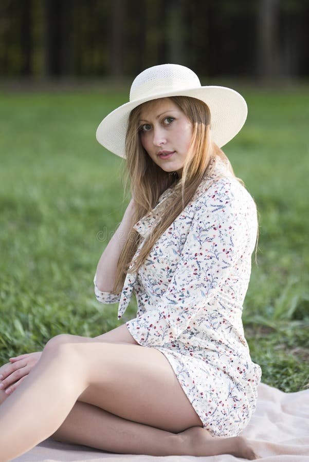 A girl in a hat in a short dress and bare feet is sitting on the grass.