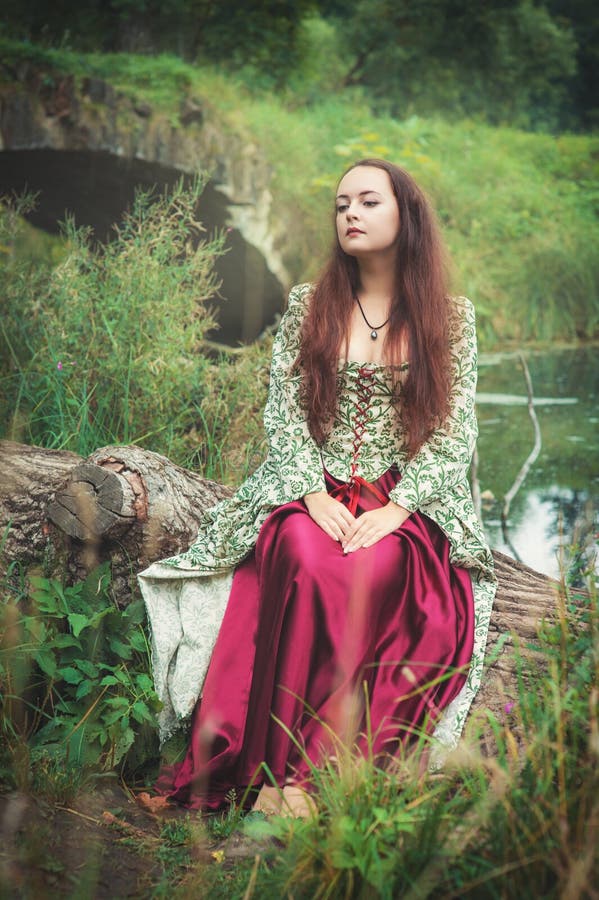 Beautiful girl in long medieval dress sitting on the tree near r