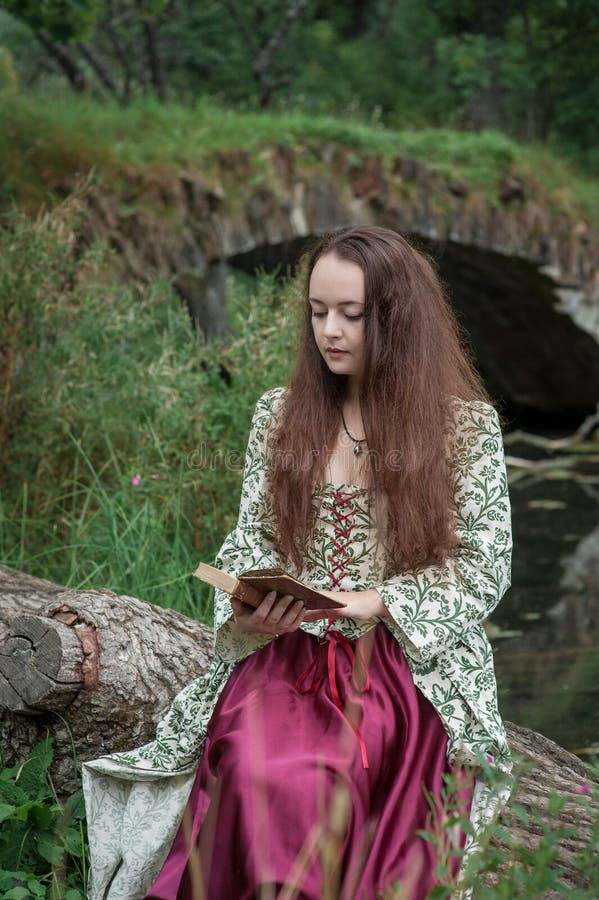 Beautiful girl in long medieval dress sitting on the tree with old book