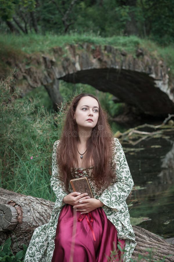 Beautiful girl in long medieval dress sitting on the tree with old book