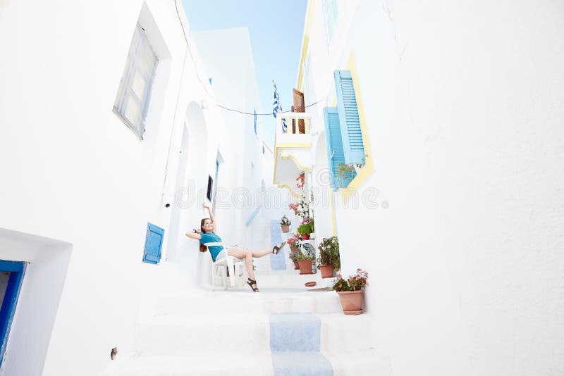 Beautiful girl on old greek street. Young attractive woman wearing shorts and sandals, sitting on chair between white traditional greek houses and enjoying stock photo Beautiful girl on old greek street. Young attractive woman wearing shorts and sandals, sitting on chair between white traditional greek houses and enjoying stock photo