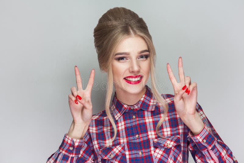 Beautiful girl with pink checkered shirt, collected updo hairstyle and makeup standing and showing victory or peace sign, looking royalty free stock image Beautiful girl with pink checkered shirt, collected updo hairstyle and makeup standing and showing victory or peace sign, looking royalty free stock image