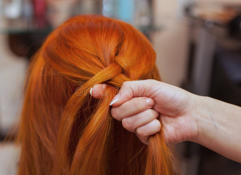 Beautiful girl with red long hair, hairdresser weaves a French braid, in a beauty salon.
