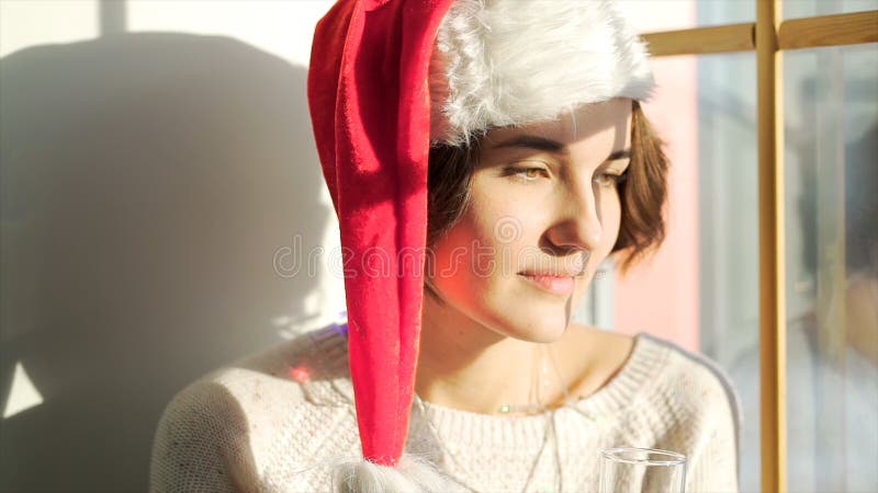 Beautiful girl in Santa Claus hat with a glass of champagne looking out of the window, Merry Christmass and New Year stock photo Beautiful girl in Santa Claus hat with a glass of champagne looking out of the window, Merry Christmass and New Year stock photo