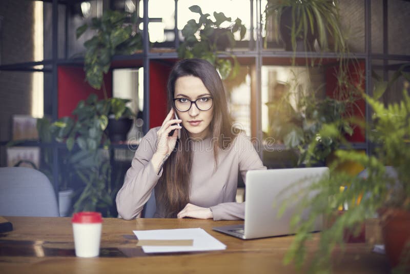 Beautiful girl wearing eye glasses in coworking studio talking by smartphone. Concept of young people working with mobile gadgets. royalty free stock image Beautiful girl wearing eye glasses in coworking studio talking by smartphone. Concept of young people working with mobile gadgets. royalty free stock image