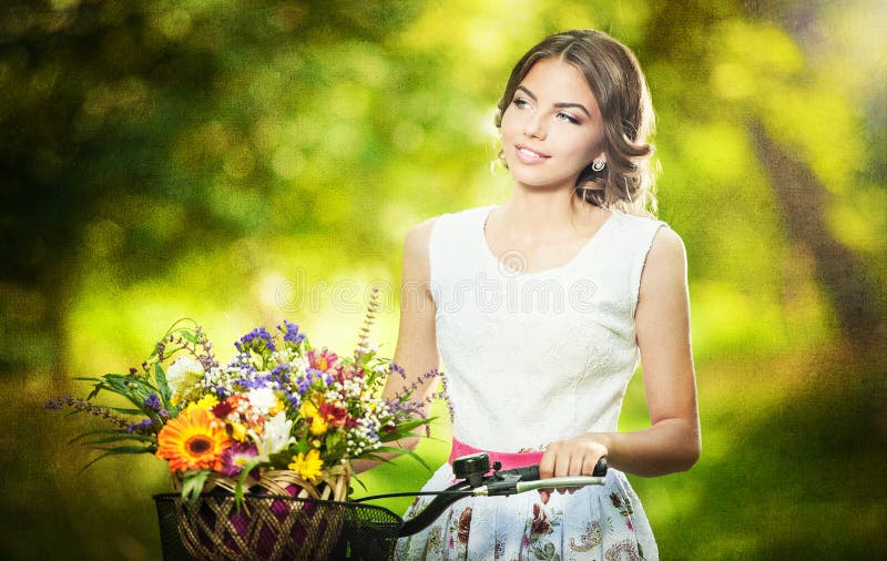 Beautiful girl wearing a nice white dress having fun in park with bicycle carrying a beautiful basket full of flowers. Vintage