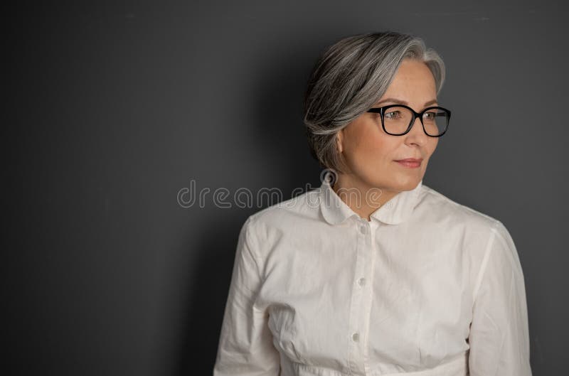 Beautiful gray-haired business woman looking at side. Mature woman in glasses and white shirt on grey background