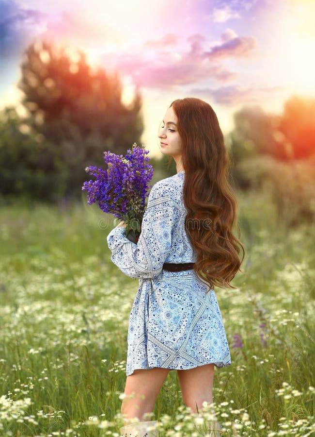 A beautiful half-turn girl in a blue dress and a summer boot standing in nature where there are wildflowers and a meadow with a bouquet of lilac lupins. Green grass and sunset background