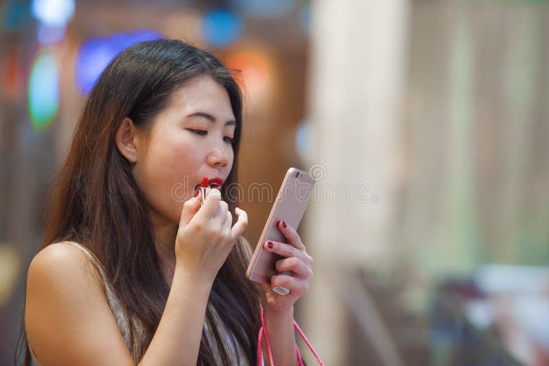 Beautiful and happy Asian Chinese woman retouching lips with lipstick makeup looking at mobile phone using it as mirror carrying s