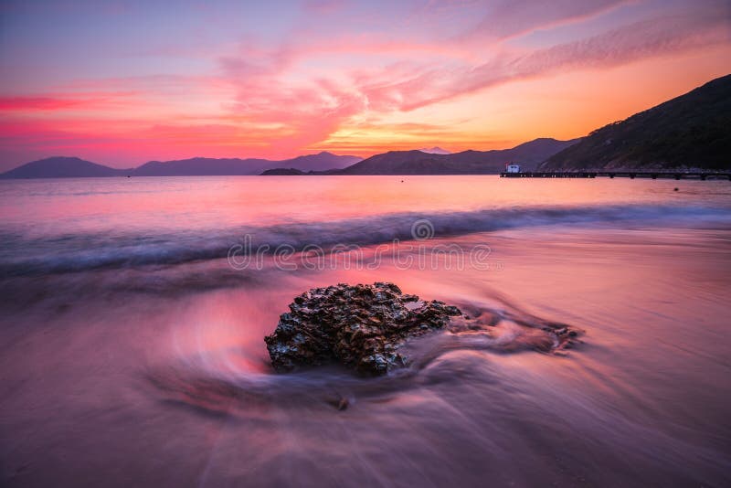 Beautiful high angle shot of a rock in a wavy sea under an orange and pink sky at sunset